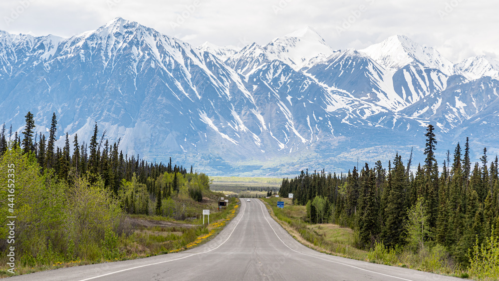 Alaska Highway leading into Haines Junction, Yukon Territory heading north west with massive snow capped mountains in the distance on cloudy, misty morning. 