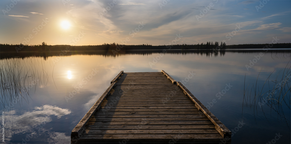 Evening sun lights up clouds and an old wooden dock in a calm lake.
