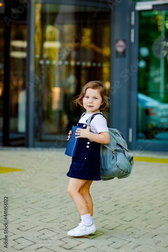 a beautiful Caucasian girl, a schoolgirl, is standing near the school, with a backpack and holding a textbook in her hands