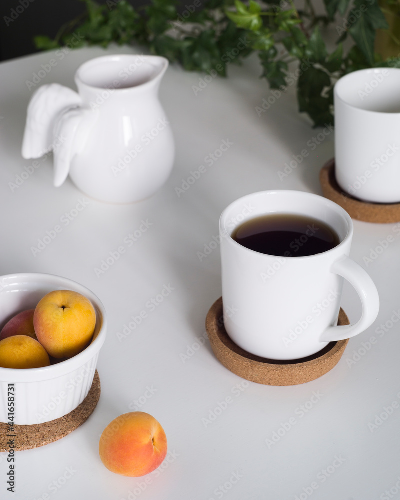 on white table, white cup with black coffee stands on brown round stand. second cup is in back out of focus. in upper left corner there is white ceramic milk jug. in lower left corner of bowl with