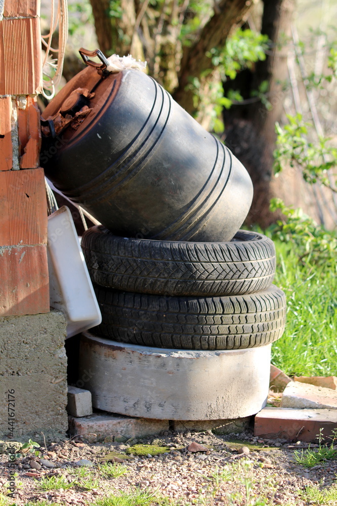Pile of junk in form of old car tyres mixed with construction material and heavily used small black plastic barrel left in suburban family house next to red brick wall on warm sunny spring day