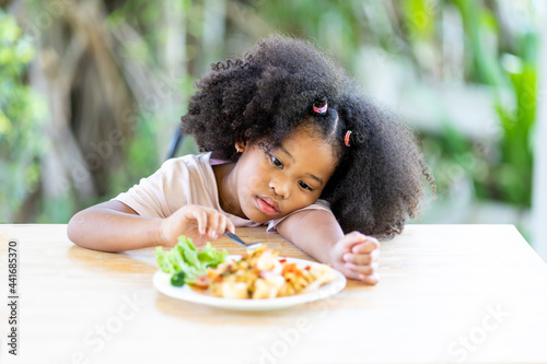 A young, curly haired African American girl sits looking at food. Childhood concepts and healthy eating.