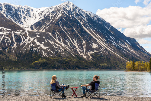 Couple sitting outdoors in mountains of Kluane National Park, Yukon Territory, northern Canada with picnic, red table between and camping, lounging chairs. Stunning blue sky day. 