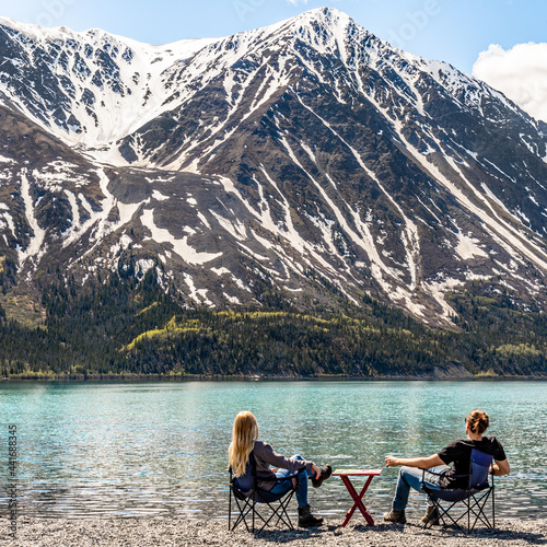 Man and woman couple sitting beside a lake in scenic mountain view setting, scene in Kluane National Park with hige snow capped mountain peaks in distance. 