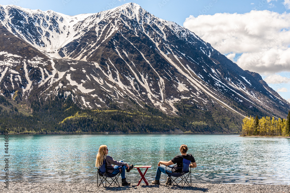 Man and woman couple sitting beside a lake in scenic mountain view setting, scene in Kluane National Park  with hige snow capped mountain peaks in distance. 