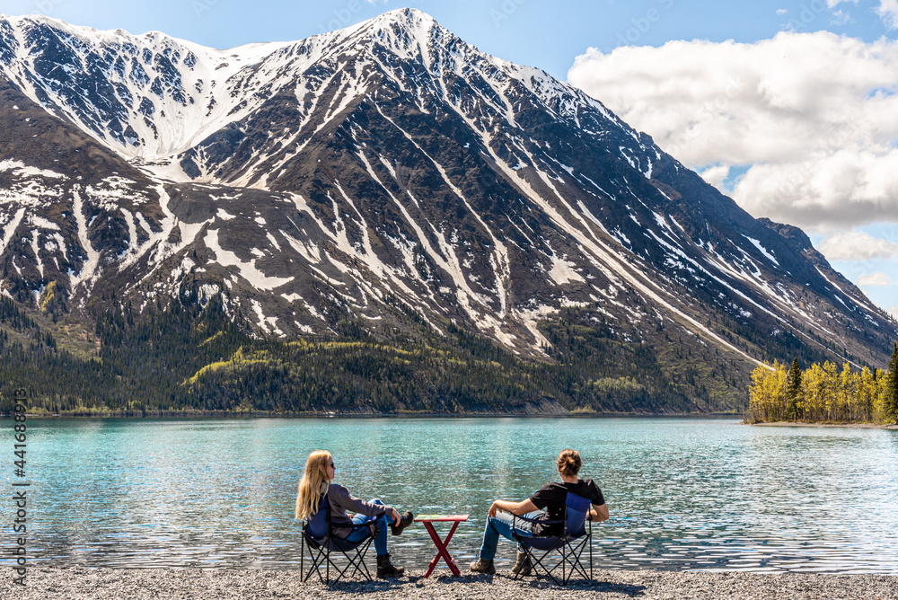 Two people, friends, couple sitting on camping chairs with picnic table on lake shore scenic area in Yukon Territory, northern Canada. Taken in early spring time on blue sky day. 