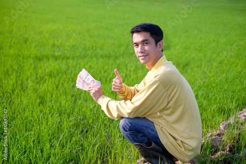 Asian farmer man wears yellow shirt sitting and holding Thai banknote money at green rice farm. photo