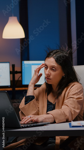 Overworked exhausted businesswoman working in startup office checking management strategy on laptop late at night. Stressed executive manager remains alone in company room after his collegue left
