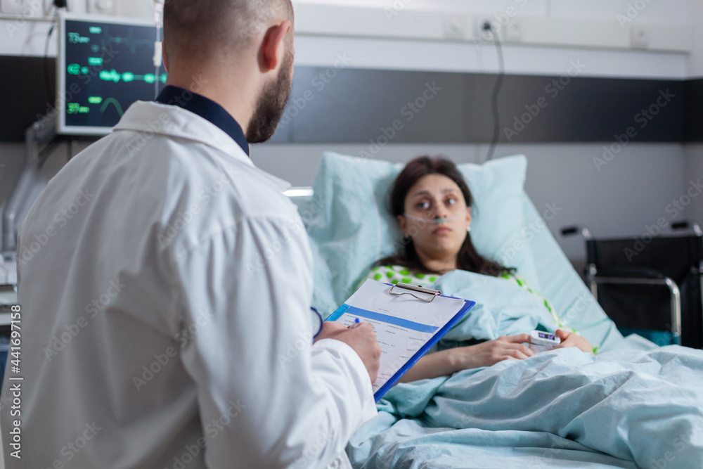 Fototapeta premium Oncologist practitioner doctor explaining sickness diagnose writing medical disease treatment on clipboard working in hospital ward. Patient sitting in bed recovering after medical surgery, health