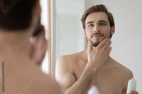 Konstfotografi Happy handsome young man applying lotion or balm on stubble after-shaving or trimming, looking at mirror, touching beard