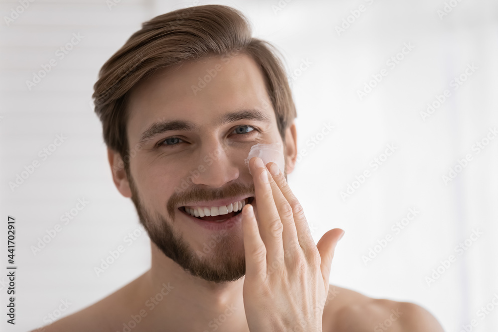 Head shot of young attractive guy applying moisturizer, sunscreen on face, applying anti age collagen cream, mask on under eye skin, preventing sun burn, wrinkles, dryness. Close up portrait Head shot of young attractive guy applying moisturizer, sunscreen on face, applying anti age collagen cream, mask on under eye skin, preventing sun burn, wrinkles, dryness. Close up portrait