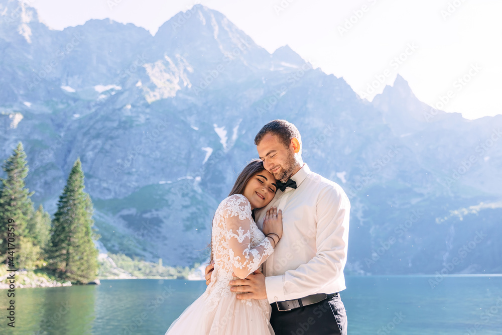 A young couple of newlyweds, the bride and groom are walking along the rocky shore of a mountain lake against the backdrop of beautiful mountains and blue sky. The bride gently hugs the man
