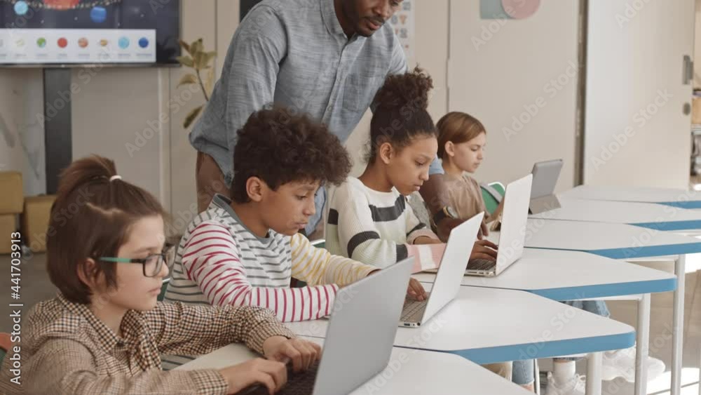 Medium slowmo of contemporary young male Afro American IT teacher helping 5 or 6 grade students working with laptops in class