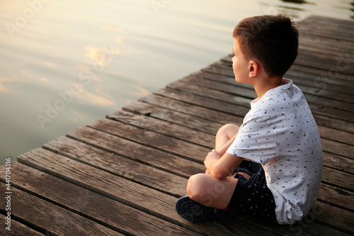 Schoolboy sitting on a pier and enjoying the tranquility of nature scene while looking at the lake landscape at sunset time