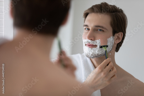 Positive attractive young guy with gel foam on lower face shaving with razor at mirror. Handsome man enjoying morning bath routine, caring for appearance, healthy facial skin. Hygiene, skincare