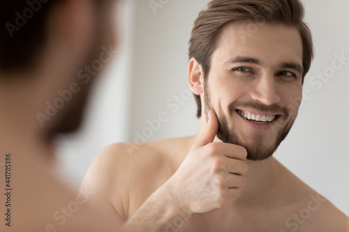 Happy millennial young handsome man looking in mirror in bathroom with toothy smile, scanning facial skin and stylish stubble. Guy satisfied with male beauty care product. Skincare, home spa concept