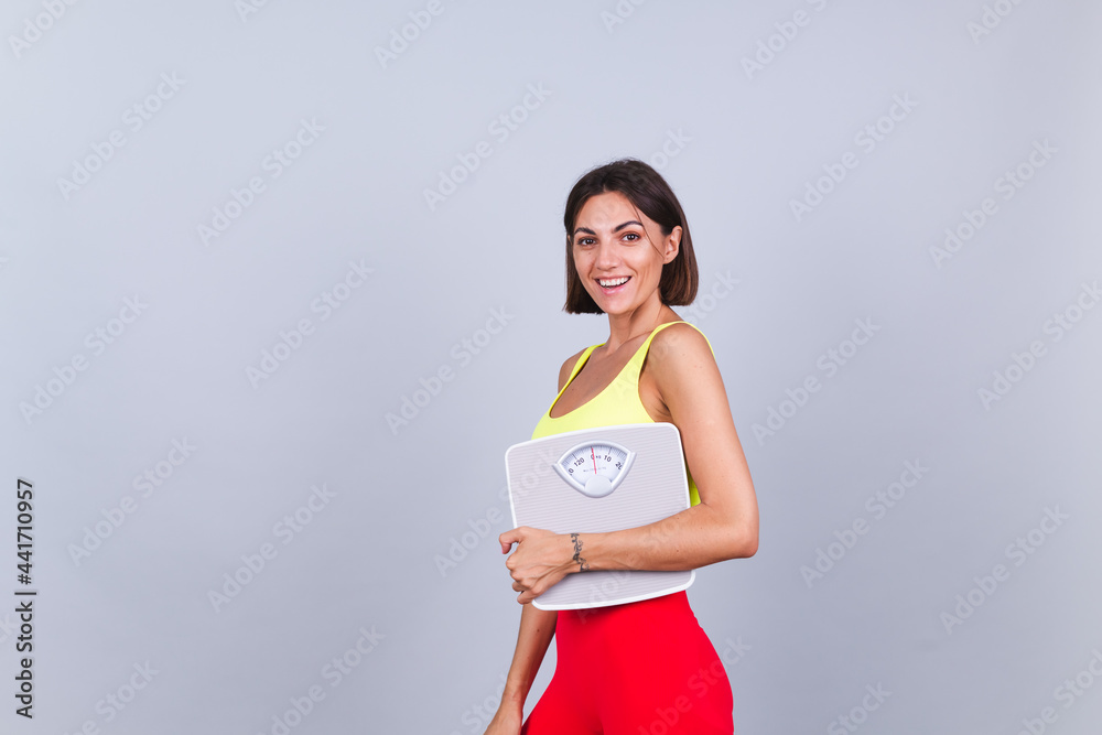 Sports woman stands on gray background, satisfied with the results of fitness training and diet, holding scale, wears top and tight leggings Sports woman stands on gray background, satisfied with the results of fitness training and diet, holding scale, wears top and tight leggings