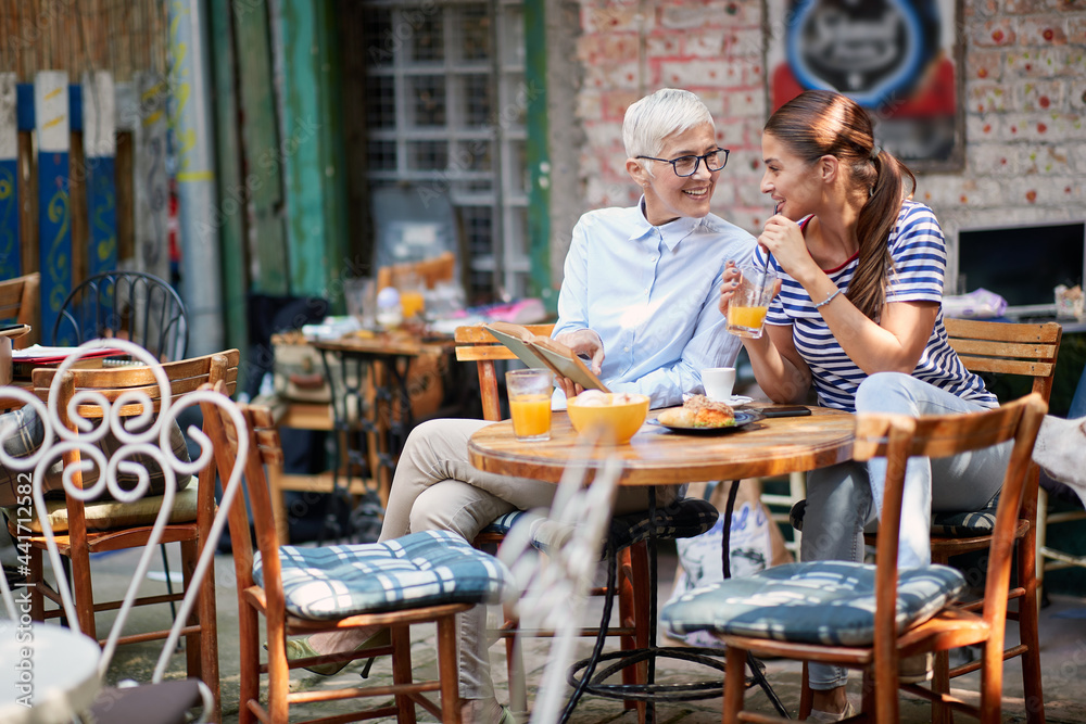 elderly woman showing part in opened book to young adult brunette female drinking orange juice on a straw, sitting in outdoor cafe