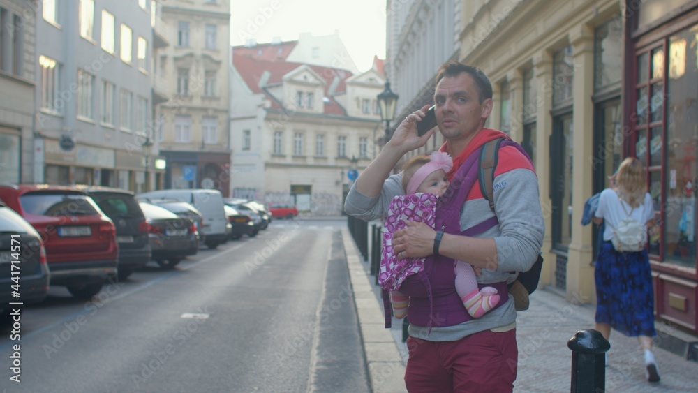 A man's dad is holding a small child in a sling. He Uses the phone, makes a call to the service. Waiting for a taxi car in the city.