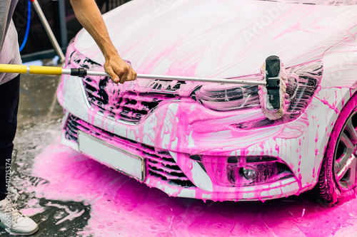 Worker washing a white car with a brush, at a car wash, a man cleans up dirt on a car with pink foam