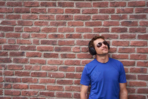 Portrait of a young man wearing headphones and a reusable mask agains coronavirus. Man staying with a face mask looking at the camera in front of brick wall with copyspace