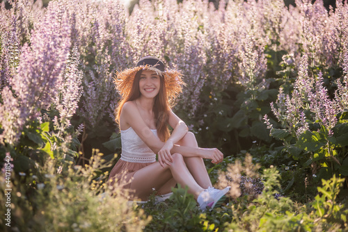 Young blonde woman in chiffon powdery jumpsuit sits against the background of blooming field of pink sage. Portrait of beautiful girlin straw hat. Weekend walk outside the city. Agricultural texture