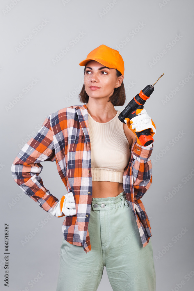 Beautiful woman in plaid shirt, hat and gloves, on gray background holds drill to repair home confident happy smile Beautiful woman in plaid shirt, hat and gloves, on gray background holds drill to repair home confident happy smile