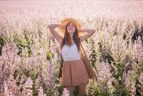 Young blonde woman in straw hat against the background of blooming field pink sage. Close-up portrait of beautiful girl holding bouquet flowers in hands in wicker basket. Agricultural texture