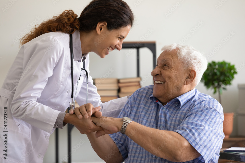 Fototapeta premium Overjoyed young Caucasian female doctor support laugh have fun with old 80s patient in clinic. Smiling woman nurse comfort caress mature elderly man client in hospital show good medical service.