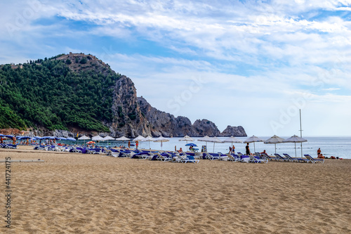 Fototapeta Naklejka Na Ścianę i Meble -  Alanya, Turkey - October 23, 2020: Umbrellas and sunbeds on the sandy Damlatas beach on the backdrop of a cliff with Alanya castle on top. Few people in a Turkish resort in the spring and autumn time