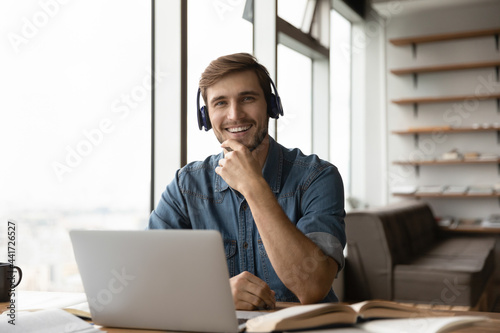 Portrait of happy millennial generation man in headphones sitting at table with books and computer. Smiling young male student posing in modern home office, e-learning distantly on online courses.