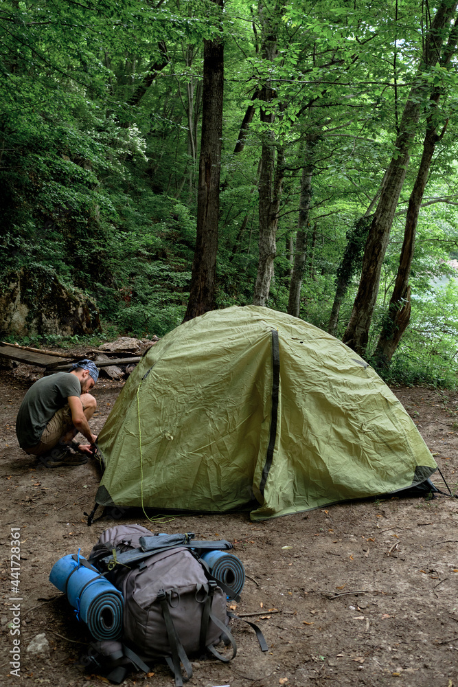 Put tourist tent in campsite and prepare for rest. Young handsome Caucasian male traveler with bandana on his head and beard sets up tent for rest in clearing in forest.