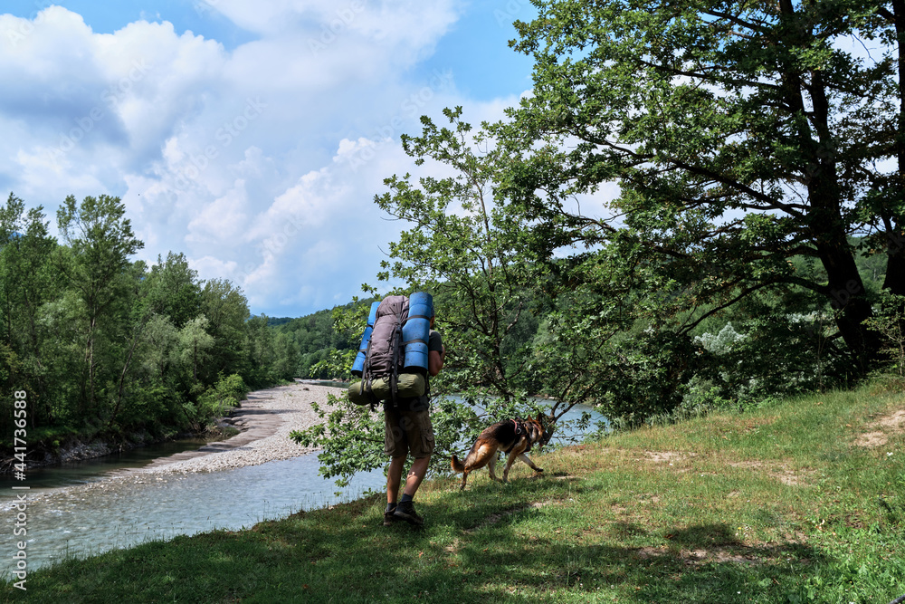 Guy is traveler and tourist. Young handsome Caucasian man with German shepherd is walking along bank of mountain river with large hiking backpack and enjoying views and beauties.