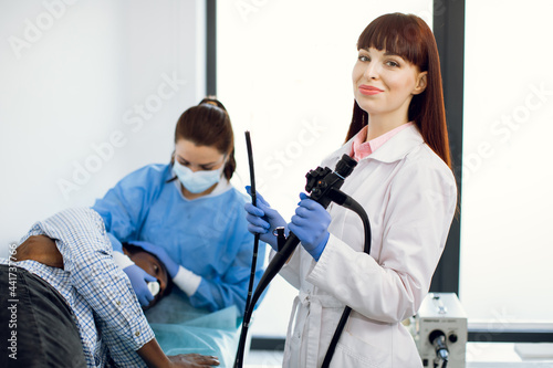 Endoscopy concept. Portrait of young Caucasian woman doctor holding endoscope before endoscopy. Nurse preparing male afro-american patient to the procedure