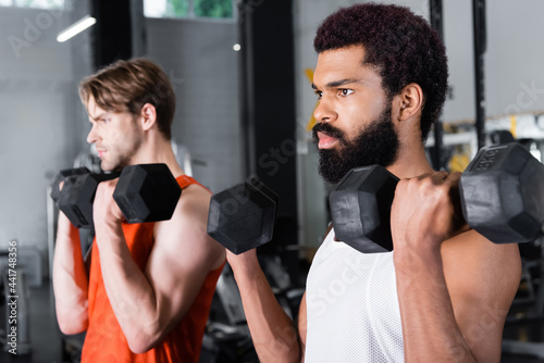 bearded african american man training with dumbbells near blurred friend