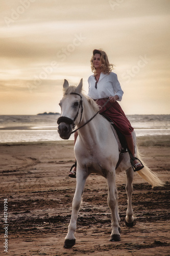 Cute happy young woman on horseback in summer beach by sea. Rider female drives her horse in nature on evening sunset light background. Concept of outdoor riding, sports and recreation. Copy space © Alex Vog