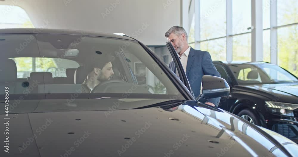 Bearded caucasian male manager giving keys from new car to happy male client. Young caucasian man sitting in auto and feeling happiness from expensive purchase. Bearded caucasian male manager giving keys from new car to happy male client. Young caucasian man sitting in auto and feeling happiness from expensive purchase.