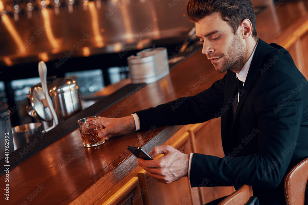 Handsome young man in full suit using smart phone while sitting at the bar counter in restaurant