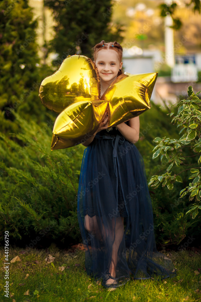 A beautiful little girl with bright yellow balloons in a blue dress stands on a green background in a city park. A holiday in nature in the summer. Children's emotions of joy.