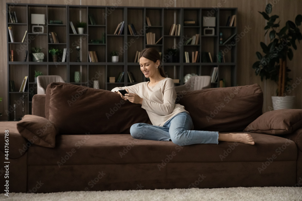 Full length happy young female homeowner relaxing on comfortable sofa, enjoying using telephone. Smiling millennial woman playing mobile game, shopping in internet store or web surfing online.