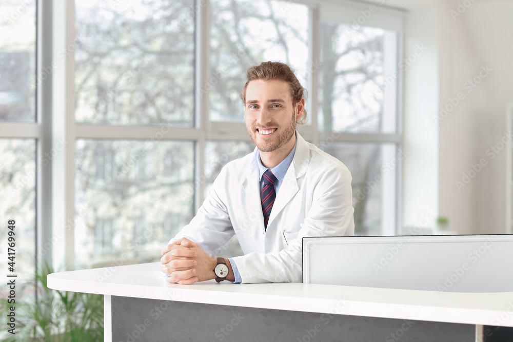 Fototapeta premium Smiling male doctor or medical professional in white lab coat at reception desk, healthcare and patient care