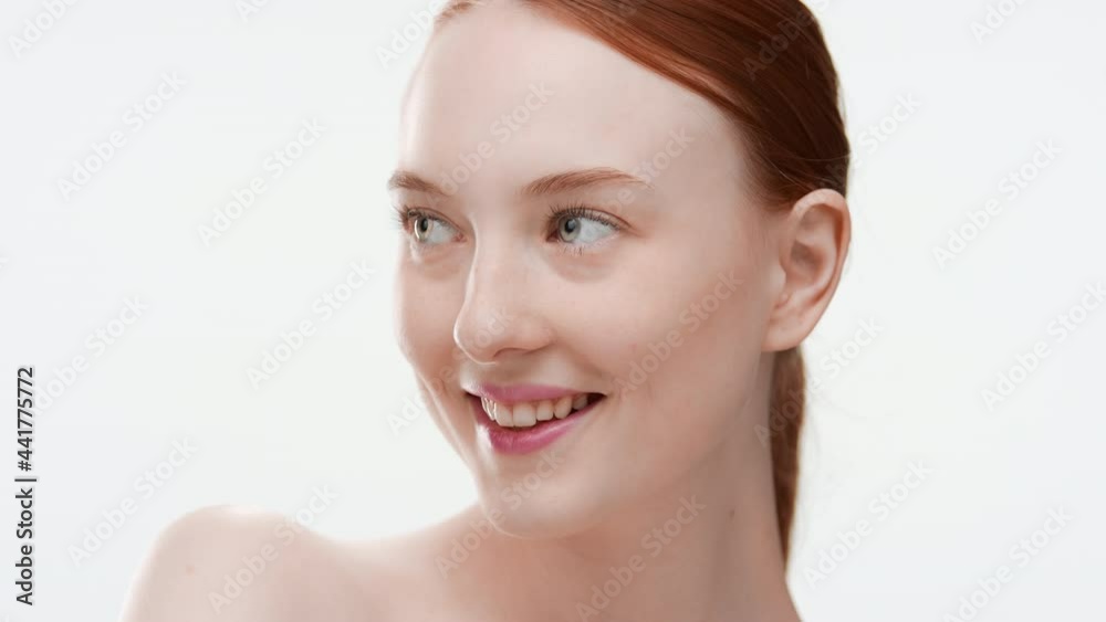 Pretty Caucasian slim young woman with long orange hair in ponytail touches her nose and cheek smiling wide for the camera on white background | Moisturizer commercial Pretty Caucasian slim young woman with long orange hair in ponytail touches her nose and cheek smiling wide for the camera on white background | Moisturizer commercial