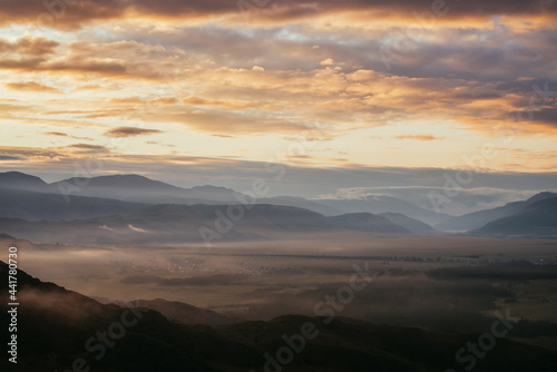 Scenic mountain landscape with golden low clouds above village among mountains silhouettes under dawn cloudy sky. Atmospheric alpine scenery of countryside in low clouds in sundown illuminating color.