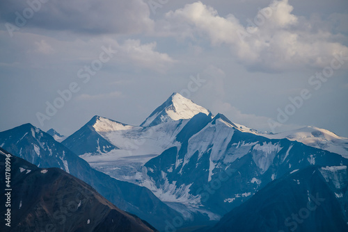 Awesome mountains landscape with big snowy mountain pinnacle in blue white colors under cloudy sky. Atmospheric highland scenery with high mountain wall with pointed top with snow under white clouds.