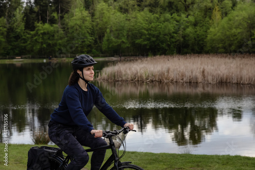 Adult Caucasian Woman Riding a Bicycle on a path by a lake in a modern city park. Spring Evening. Taken in Green Timbers Urban Forest, Surrey, Vancouver, British Columbia, Canada.