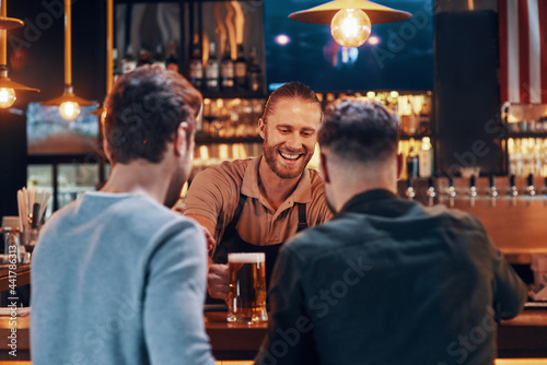 Cheerful bartender serving beer to young men while standing at the bar counter in pub