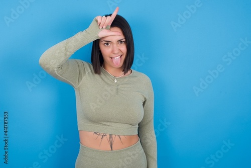 Funny Young beautiful sporty girl standing against blue background makes loser gesture mocking at someone sticks out tongue making grimace face. photo