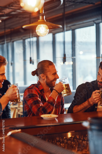 Happy young men in casual clothing enjoying beer and smiling while sitting at the bar counter in pub