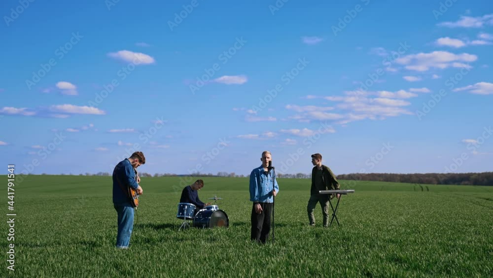 Outdoor concert of young musicians. Group of friends playing the guitar, drums and synthesizer performing music on green field under blue sky. Outdoor concert of young musicians. Group of friends playing the guitar, drums and synthesizer performing music on green field under blue sky.
