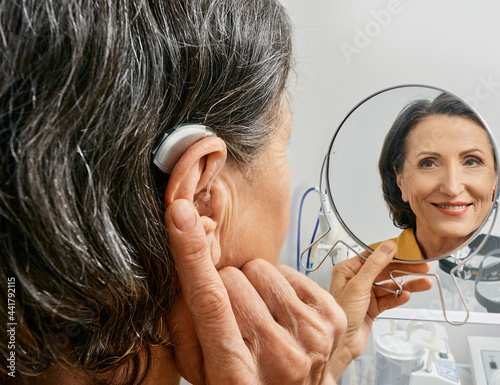 Fotografie Mature woman with BTE hearing aid looks at herself in mirror and tries on hearing device, close-up